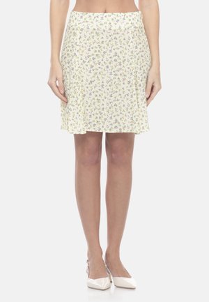 Femme portant une jupe crème mi-longue avec un petit imprimé floral violet et jaune et des talons blancs pointus.