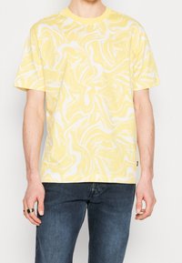 Man i en kortärmad t-shirt med gul och vit marmorering och en liten "BOSS"-logotyp på bröstet, tillsammans med mörkblåjeans.