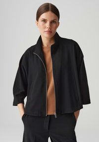 Black zip-up jacket with a high collar, relaxed fit, and three-quarter sleeves, paired with a brown shirt and black pants. Smooth fabric texture.