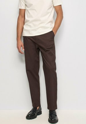 Hombre con pantalones rectos marrón oscuro, zapatos negros y una camisa de manga corta color crema, con una mano en el bolsillo, de pie frente a un fondo liso.