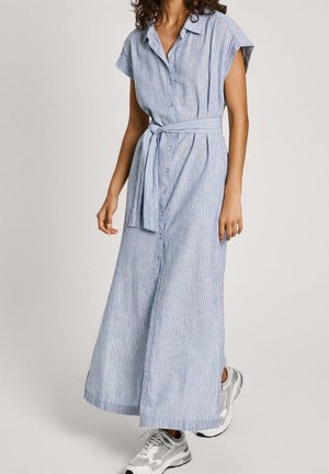 Kobieta ubrana w długą sukienkę w pionowe paski w jasnoniebieskim i białym kolorze, z krótkimi rękawami, zapinana na guziki z przodu, z paskiem w talii i białymi trampkami.