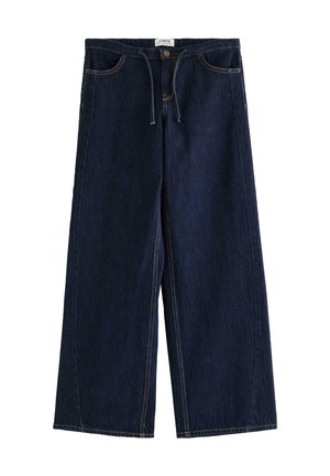 Pige stående iført en hvid kortærmet t-shirt, vidde mørke jeans og hvide sneakers mod en ensartet lys baggrund.