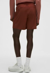 Bărbat îmbrăcat în pantaloni scurți sport de culoare ruginie, tricou lung cu mâneci roz, sosete albe și pantofi de alergare albi, stând în fața unui fundal simplu.