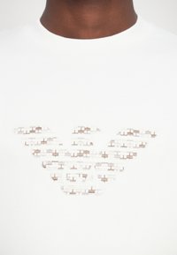 Valge puuvillane T-särk, millel on suur, tikandiga logo pehmes beeži ja halli toonis, tiibade kuju ning keerukalt kudumiskonstruktsioon.