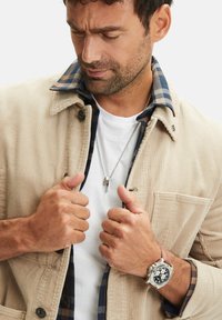 Lichte beige corduroy jas met een geruite blauwe en zwarte kraag, gedragen over een wit t-shirt. Zilveren horloge om de pols.