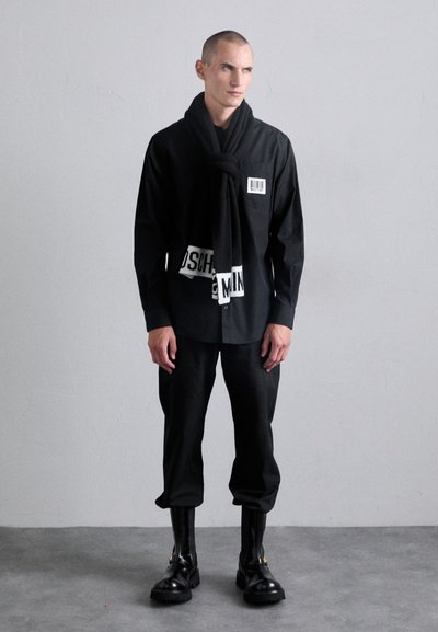 Svart skjorte med knapper og strekkode-tag, kombinert med svarte bukser og kraftige svarte støvler. Outfitet inkluderer et knyttet svart skjerf med hvite detaljer.