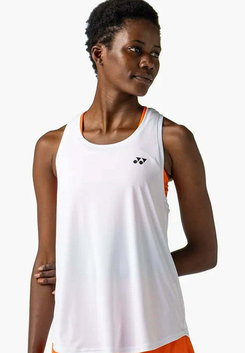 Sportliche Frau, die ein locker sitzendes weißes ärmelloses Oberteil über einem orangen Sport-BH trägt, schaut nach rechts und hat einen Arm hinter dem Rücken.