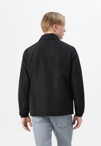 Veste légère noire avec un col, des poignets élastiques et une coupe décontractée. Portée sur un jean gris clair, l'arrière est sans ornement et lisse.