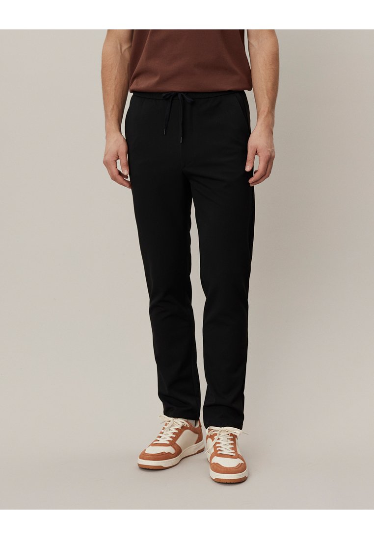 Schwarze Stretch-Hosen mit einem Kordelzugbund, schmalem Schnitt und seitlichen Taschen, kombiniert mit braunen und weißen Sneakern.