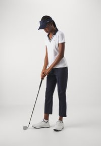 Bílá polo tričko s námořnickými akcenty, námořnické zkrácené kalhoty a bílé golfové boty z kompozitního materiálu. Drží golfovou hůl, má na sobě námořnickou kšiltovku.