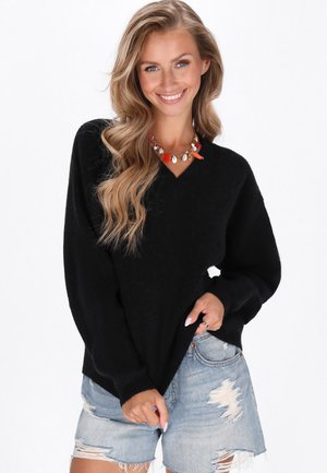 Donna sorridente con lunghi capelli mossi indossa un maglione nero a scollo a V e pantaloni shorts in denim strappati, accessoriata con una collana colorata.