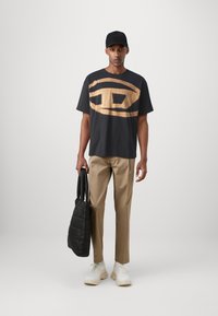 Sort t-shirt med et fremtrædende lysebrunt logo, beige bukser, hvide sneakers og en sort quiltet totebag. Enkel, afslappet påklædning.