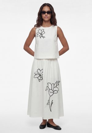 Donna con top corto bianco senza maniche e gonna lunga con disegni floreali neri a linea, ballerine nere e occhiali da sole neri stretti, in piedi immobile.