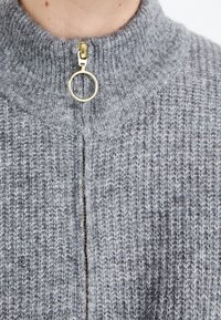 Maglione grigio lavorato a maglia con colletto alto e una cerniera dorata con un tirante circolare. Il tessuto presenta una texture a coste e una leggera variazione di colore.