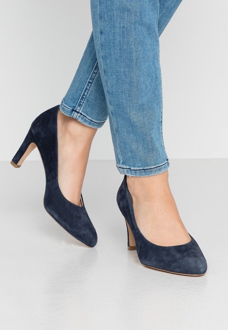 Tummansininen mokkanahkainen korkokenkä, jossa on pyöreä kärki ja pieni vaaleanruskea paksu korko, yhdistettynä nilkkapituisiin vaaleansinisiin farkkuihin.