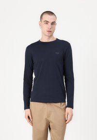 T-shirt a maniche lunghe blu navy in cotone, con colletto a girocollo e un piccolo logo stampato sul petto. Abbinato a pantaloni khaki.