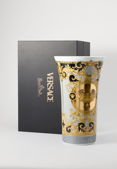 Vaso de cerâmica com padrão barroco em azul e dourado, apresentando motivos de leão e detalhes luxuosos, ao lado de uma caixa de presente preta da Versace.