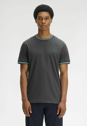 Junger Mann mit kurzem Afro-Haarschnitt, der ein dunkelgraues T-Shirt mit grün-weißen Streifen am Kragen und an den Ärmelsäumen trägt, steht aufrecht.