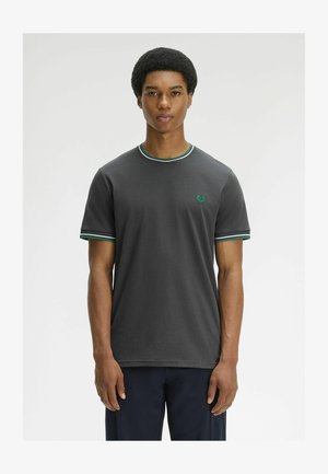 Jonge man met korte afro-haarstijl draagt een donkergrijs T-shirt met groene en witte gestreepte kraag- en mouwranden, staand.