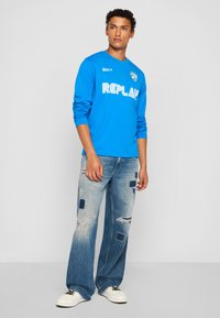 Ung mand iført en lys blå langærmet skjorte med "REPLAY" tekst og lapperede slidte blå jeans, stående mod en ensfarvet baggrund.