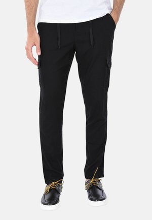 Uomo con pantaloni cargo neri con tasche laterali, camicia bianca e sneakers nere con lacci gialli su sfondo bianco.