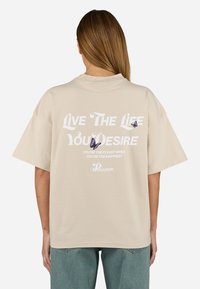 T-shirt di cotone beige con maniche corte, caratterizzata da testo bianco e grafiche di farfalle viola sul retro. Vestibilità rilassata e design semplice.