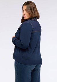 Cardigan de culoare albastru marin cu o emblema din denim. Material moale cu textură, închidere cu nasturi, manșete și tiv striate. Croială casual, detalii de cusătură vizibile.
