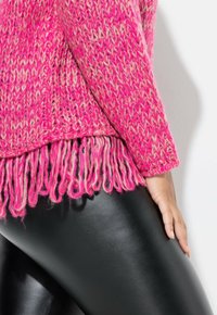 Fuchsienfarbener Strickpullover mit Chevron-Muster, der am Saum lange Fransen aufweist. Kombiniert mit glänzenden schwarzen Leggings.