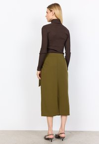 Brauner gerippter Rollkragensweater kombiniert mit einem olivegrünen Midi-Rock mit hoher Taille. Die Schuhe weisen schwarz-weiße gestrichelte Akzente auf.