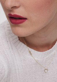 Złoty naszyjnik w kształcie nieskończoności i serca, delikatny łańcuszek, noszony na fakturowanym białym swetrze. Zbliżenie ust z różową pomadką.