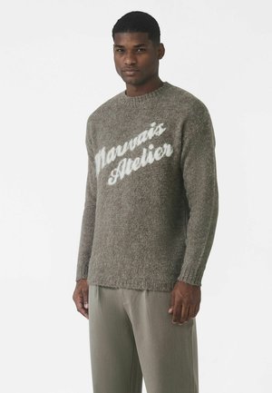 Szary sweter dziergany o puszystej fakturze, z napisem "Marvais Atelier" w białych literach na przodzie, zestawiony z jasnymi spodniami.