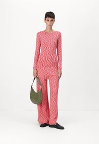 Red and white patterned jumpsuit with long sleeves and wide legs, paired with an olive green shoulder bag. Black shoes complete the outfit.