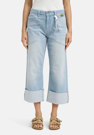 Light blue wide-leg cropped jeans with large front pockets, cuffed hems, and white tag detail, worn with beige buckle sandals.