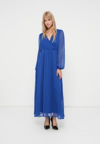 Mulher vestindo um vestido maxi azul royal de manga longa e transparente, com corpete envolvente e sapatos de salto alto pretos, de pé contra um fundo branco liso.