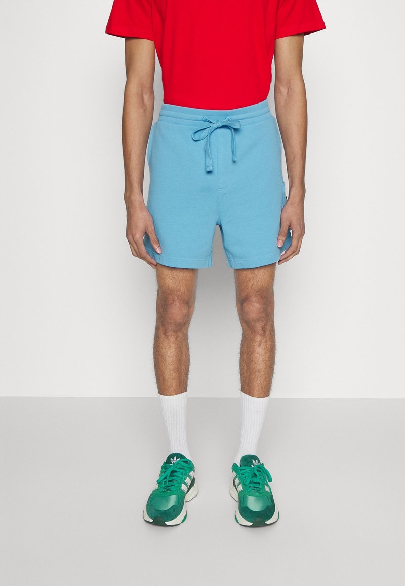 Pantalones cortos azules de algodón con cintura ajustable, combinados con una camiseta roja, calcetines blancos y zapatillas deportivas verdes.