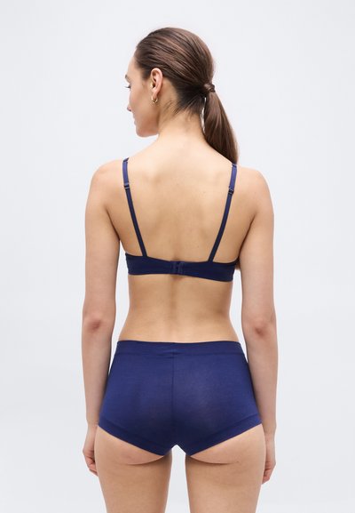 Γυναίκα με καστανά μαλλιά σε χαμηλή αλογοουρά, που φοράει μπλε ναυτικό σουτιέν και ψηλόμεσο σορτς, εμφανιζόμενη από πίσω σε λευκό φόντο.