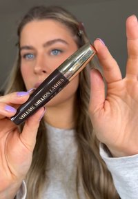 Tube de mascara noir avec des accents dorés, étiqueté "VOLUME MILLION LASHES." Tenue par une personne avec des ongles violet clair, sur un fond neutre.