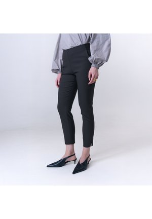 Person trägt schwarze, schmal geschnittene Knöchelhosen, eine graue Bluse mit langen Ärmeln und gerafften Manschetten sowie schwarze Slingback-Pumps mit spitzer Schuhspitze.