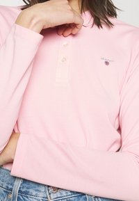 Långärmad rosa polo skjorta med krage, tre knappar och en liten logotyp med ett sköldmönster på vänster bröst. Texturerad tyg.