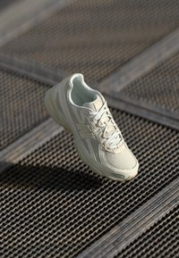 Scarpa da corsa beige chiaro con tomaia in rete, lacci leggeri e suola ammortizzata, mostrata su una superficie metallica texture.