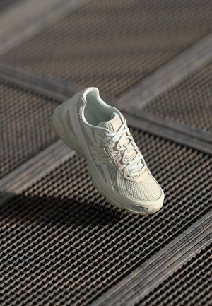 Lys beige løbesko med en mesh-overdel, lyse snørebånd og polstret sål, præsenteret mod en struktureret metalsurface.