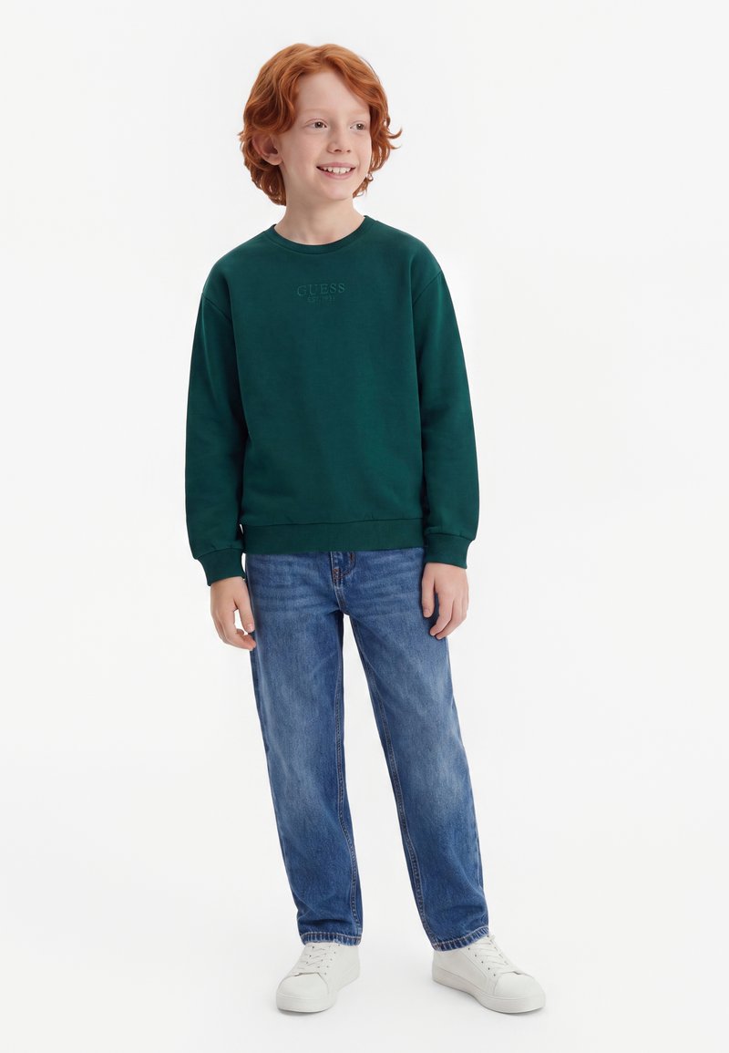 Garçon roux souriant, portant un sweat-shirt vert foncé, un jean bleu et des baskets blanches, debout devant un fond uni.
