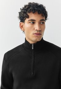 Zwarte trui met ritssluiting, voorzien van een hoge kraag, geribbelde textuur en lange mouwen met subtiele naaddetails. Gladde stofafwerking.