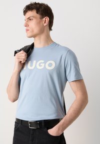 Világoskék pamut póló fehér "HUGO" felirattal, kerek neckline-nel, fekete farmernadrággal és egy szürke dzsekivel, ami az egyik vállán van. 