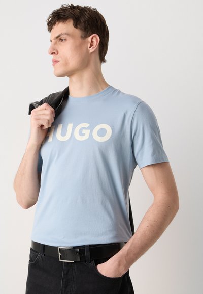 Светлосиня памучна тениска с бели букви "HUGO", кръгло деколте, носена с черни дънки и сива якет, спусната на едно рамо.