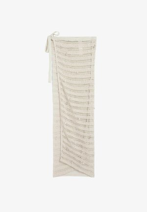 Pikk beež avatud silmusega heegeldatud seelik, millel on ümberlõikega disain ja kinnitatud vöösidemed, paigutatud valgele taustale.