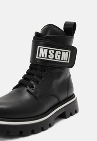 Botte de cheville en cuir noir avec une semelle épaisse, un avant lacé et une bande de logo blanche bien visible. Elle présente une finition texturée et un col rembourré.