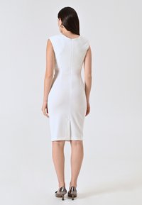 Weißes, figurbetontes Kleid mit kurzen Ärmeln, nahtlosem Design, Reißverschluss hinten in der Mitte und knielangem Saum. Getragen mit metallischen High Heels.