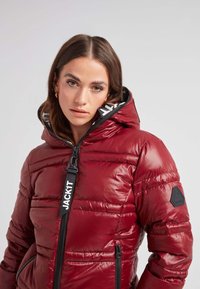 Glansig röd puffjacka med dragkedja fram, svarta detaljer, logotypaccenter och en huva med en kontrasterande innersida.