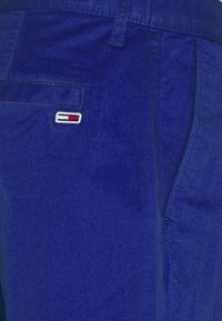 Detailní záběr na modré kalhoty ukazující zadní kapsu s malým vyšitým logem, které obsahuje červené, bílé a černé vodorovné pruhy.